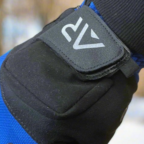 Close-up of a gloved hand gripping a bicycle handlebar. The Black & Blue GEN2 Heated Sports Glove from arcticreaction, with touchscreen compatibility, features a logo. The gear shifter displays 7 against a snowy landscape with blurred trees.