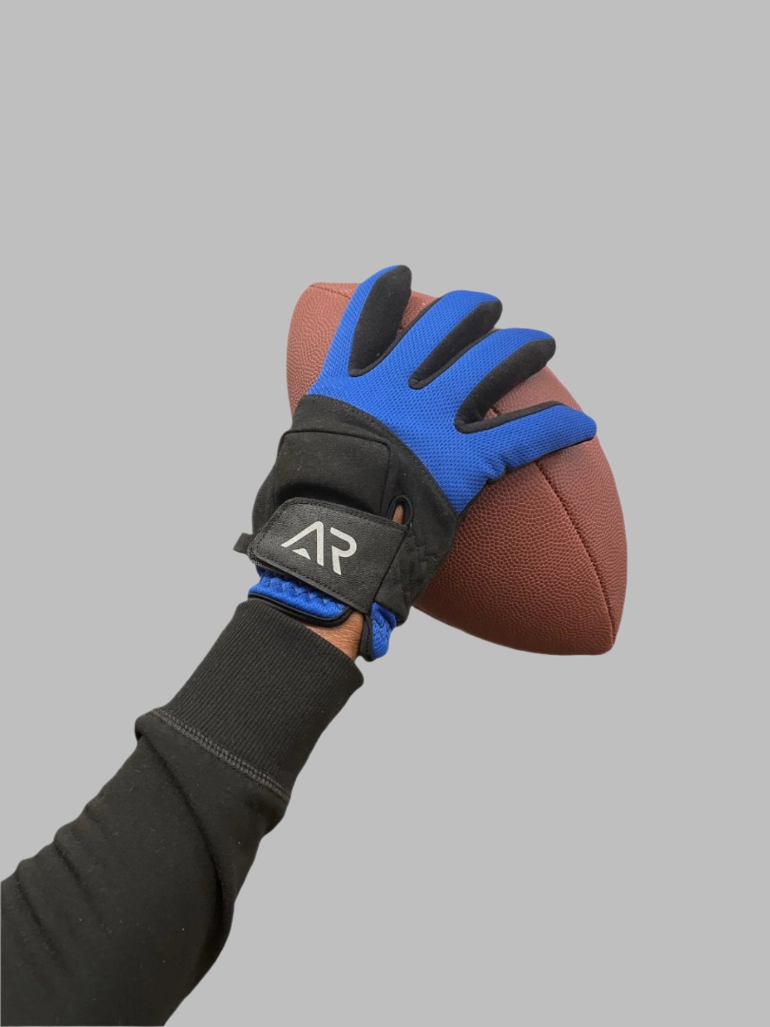 A person wearing a black sleeve and the arcticreaction Black & Blue GEN2 Heated Sports Glove is holding a brown football against a plain gray background.