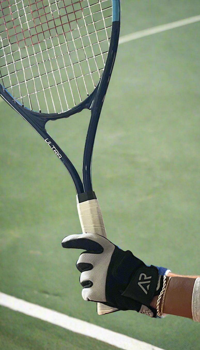 A person wearing an arcticreaction Black & Gray GEN2 Heated Sports Glove, compatible with touchscreens, holds a blue tennis racket with white strings. The racket displays the word "Ultra" on its side. In the background, there is a green tennis court outlined by a white line.