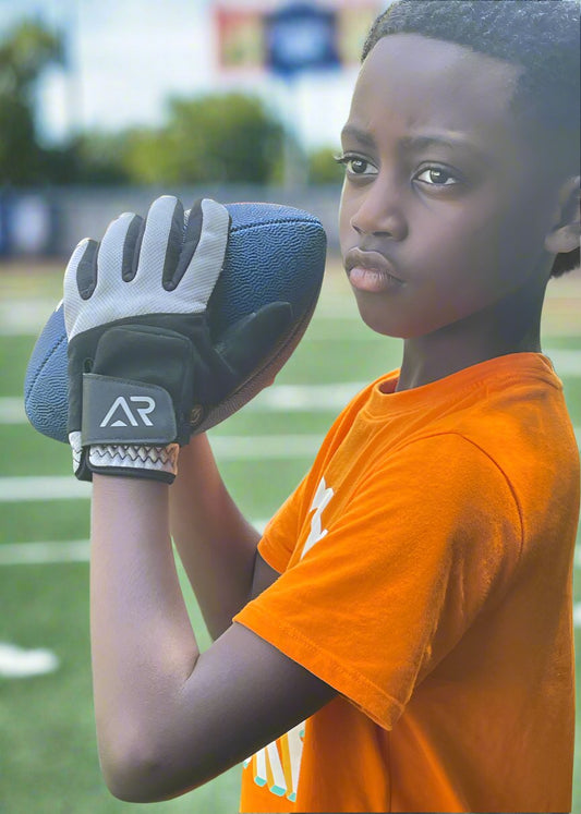 A young person stands on a football field, holding a football and wearing a bright orange shirt. They look focused and determined, with one hand clad in an arcticreaction Black & Gray GEN2 Heated Sports Glove with touchscreen compatibility, as stadium seats blur in the background.