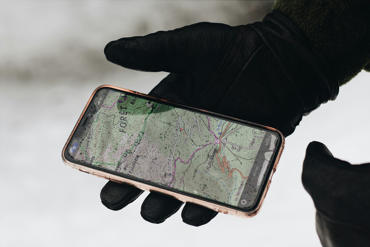 A person wearing arcticreaction's Black & Gray GEN2 Heated Sports Glove, featuring touchscreen compatibility, holds a smartphone displaying a detailed topographic map with contour lines and trails. The blurred background implies an outdoor setting, possibly snowy or misty, ideal for exploring with the gloves' dual heat settings.